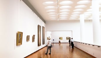 A spacious museum hall with visitors looking at various paintings on the wall.